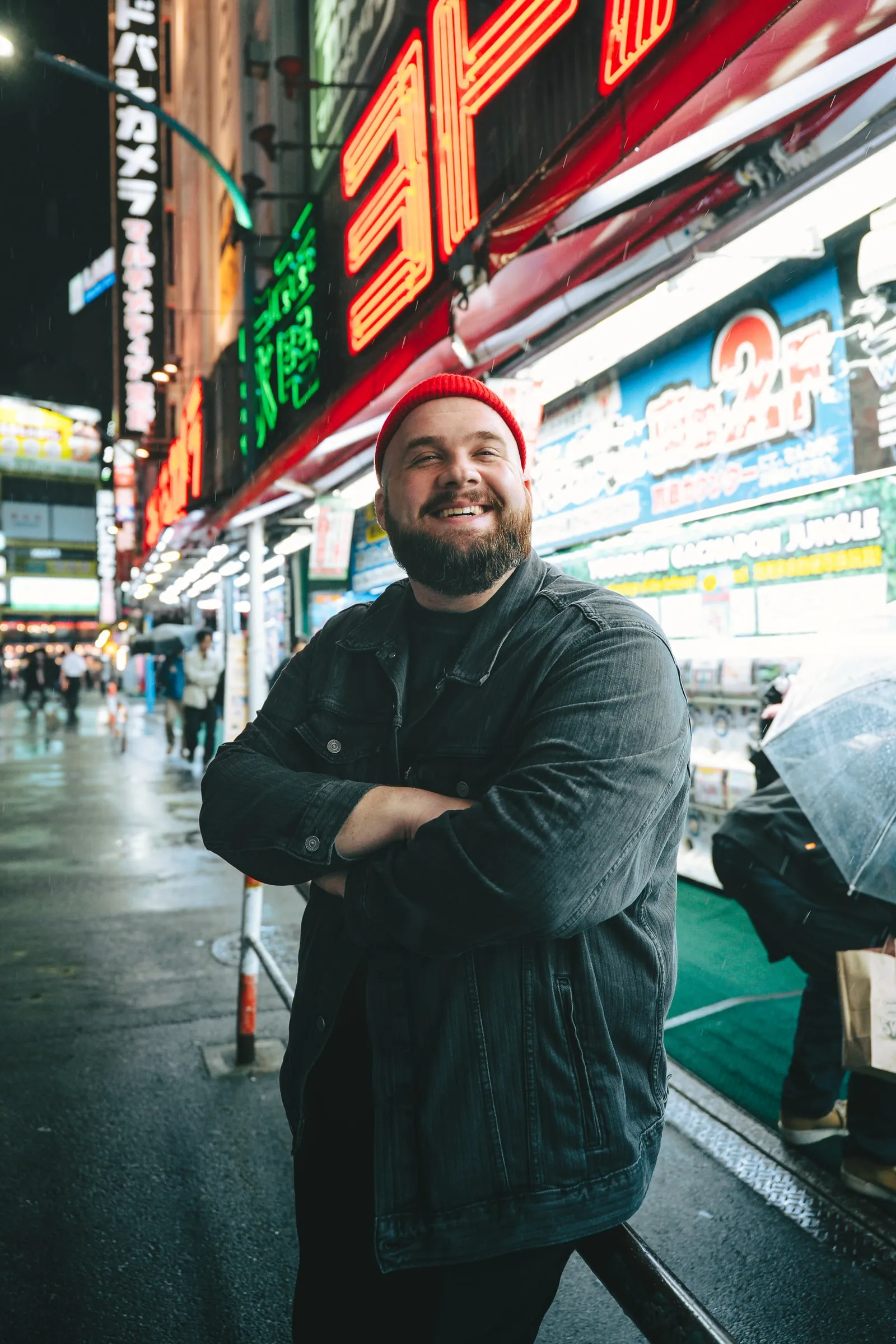 Chris Hawk in a red beanie and denim jacket, arms crossed and smiling under neon signs in Shinjuku, Tokyo.